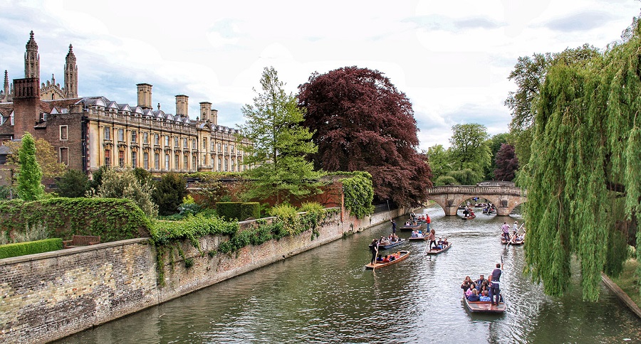 A collection of boats with people punting along the River Cam with bridges in background.