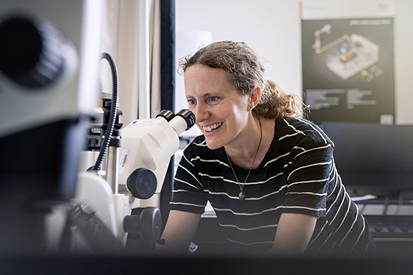 Dr Sarah Robinson looking down eyepiece of a microscope in the SLCU Microscopy Facility. Sarah is smiling.