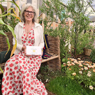 Melanie Sadler sitting on willow bench in The Pollinator Patch garden with sunset coloured calendulas in foreground and twisted willow tree and willow bicycle in background