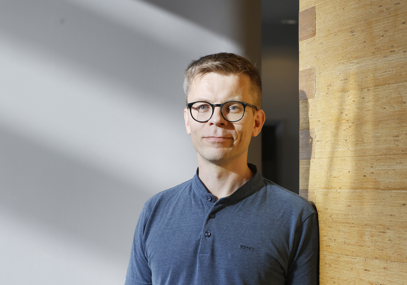 Professor Ari Pekka Mähönen standing indoors beside a wooden wall, wearing glasses and a blue shirt, with sunlight casting soft shadows across the background.