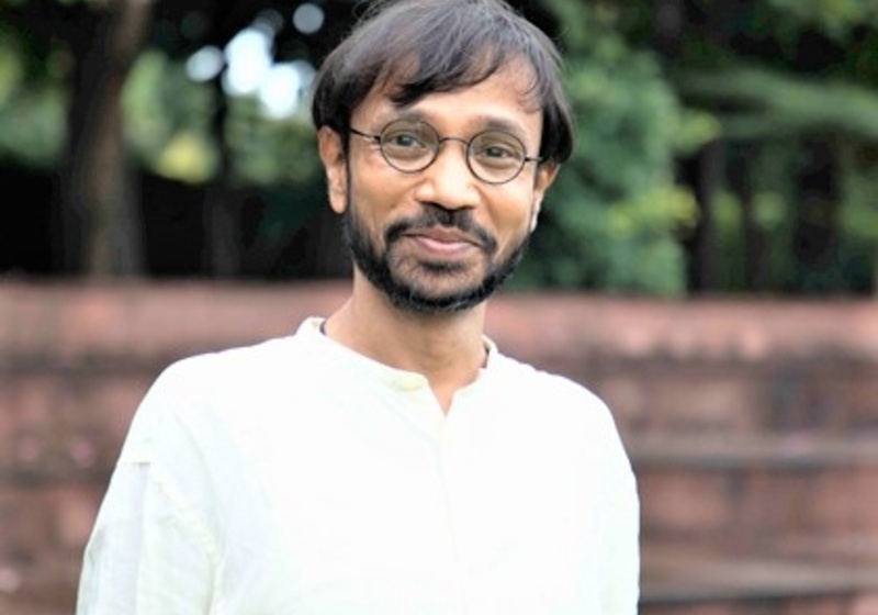 Professor Kalika Prasad standing outdoors with fence and trees in background, wearing white top and glasses