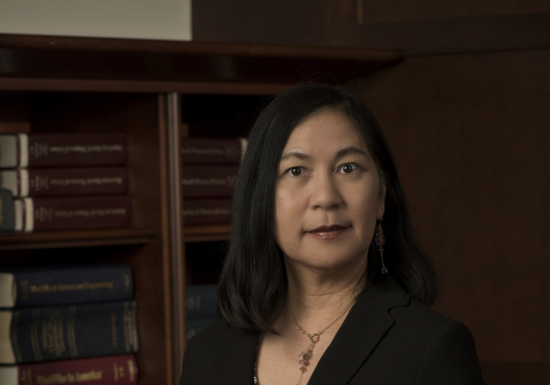 Professor Keiko Torii standing indoors with dark timber bookcase with large texts in background, wearing a black jacket