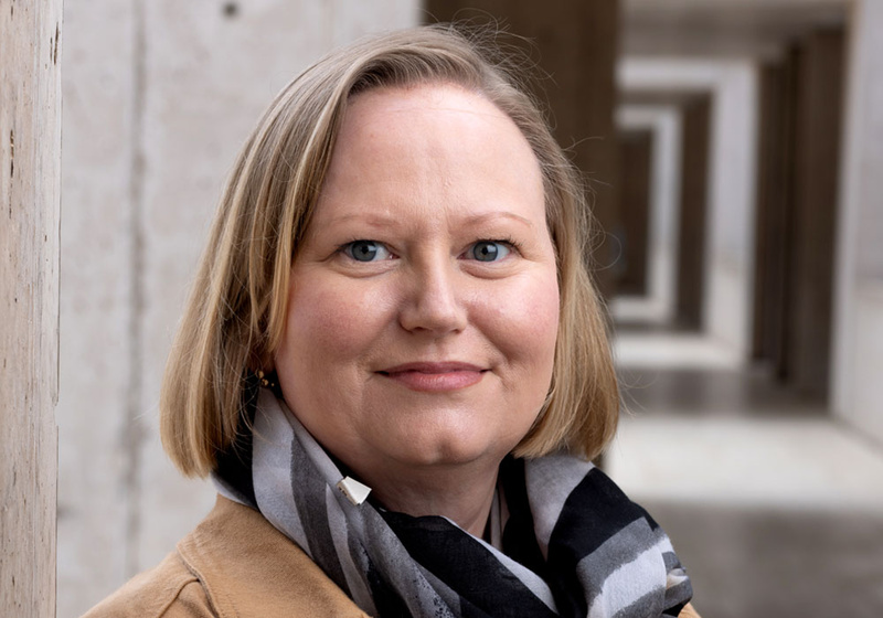 Lucia Strader standing indoors beside grey concrete wall with hallway in background, wearing a brown jacket and black and grey scarf