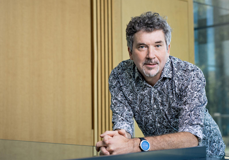 Professor Henrik Jönsson leaning forward resting elbows on shelf with oak timber and glass of internal Sainsbury Laboratory building in background