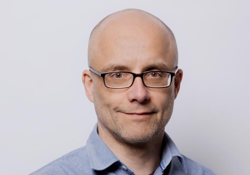 Thomas Greb standing indoors in front of white wall, wearing a blue shirt and glasses