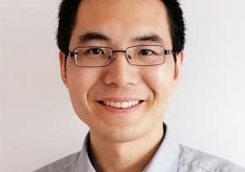 Weibing Yang standing indoors with white wall background, wearing a grey shirt and glasses