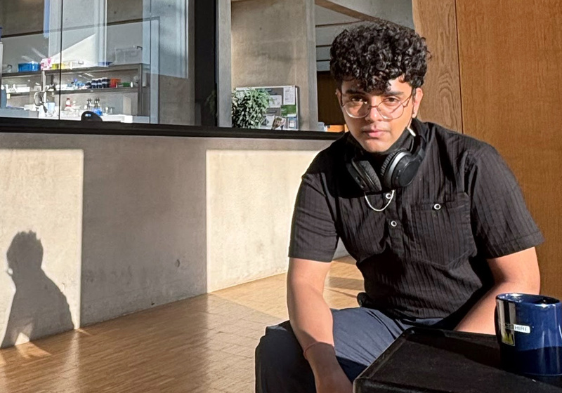 Chetan Pandey sits on a chair inside the Sainsbury Laboratory building, sunlight illuminating Chetan's face and casting shadow of Chetan on the concrete wall behind. The experimental laboratories are visible in the background.