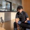 Chetan Pandey sits on a chair inside the Sainsbury Laboratory building, sunlight illuminating Chetan's face and casting shadow of Chetan on the concrete wall behind. The experimental laboratories are visible in the background.